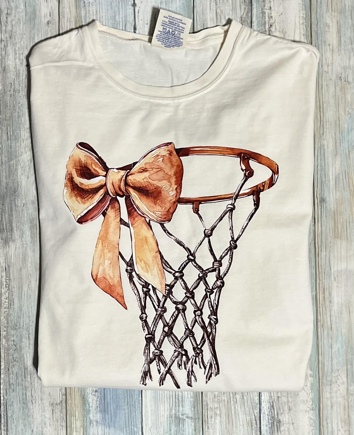 A off white long-sleeve basketball tee with a bow and net basketball hoop design, displayed on a wooden background.