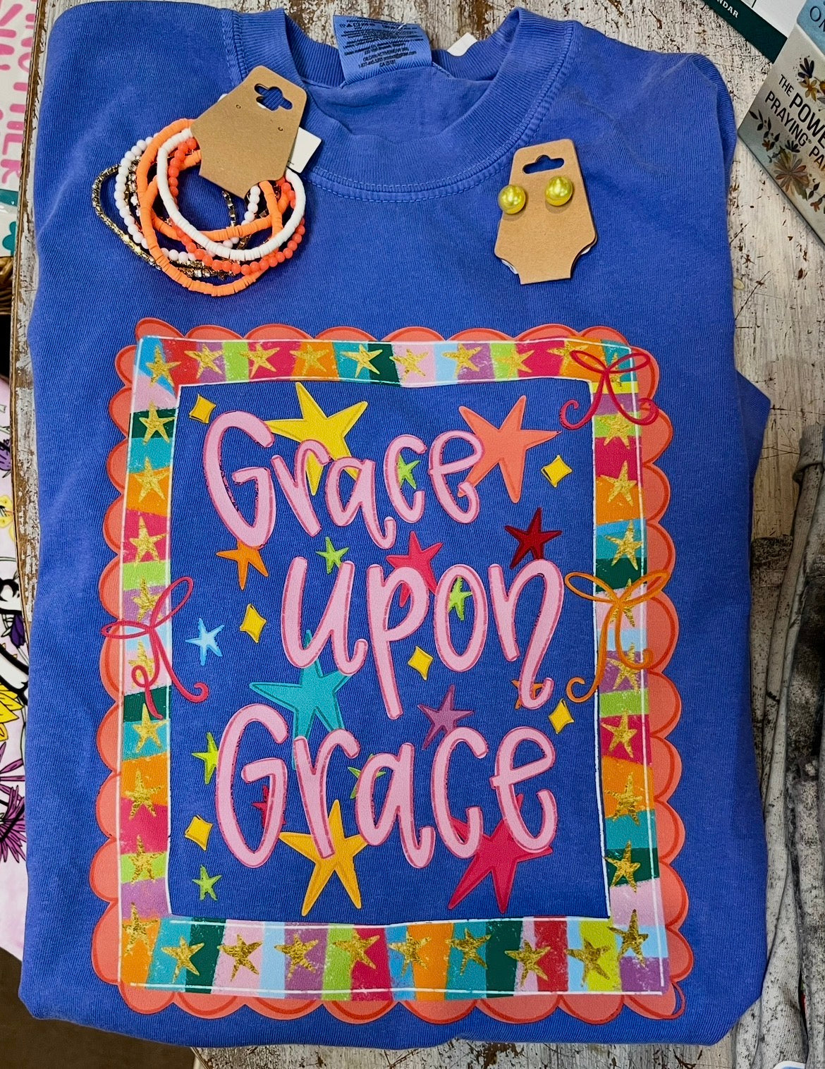 blue short sleeve tee with colorful decal that says grace upon grace on wooden background orange and white bracelet and yellow earrings laying on shirt.