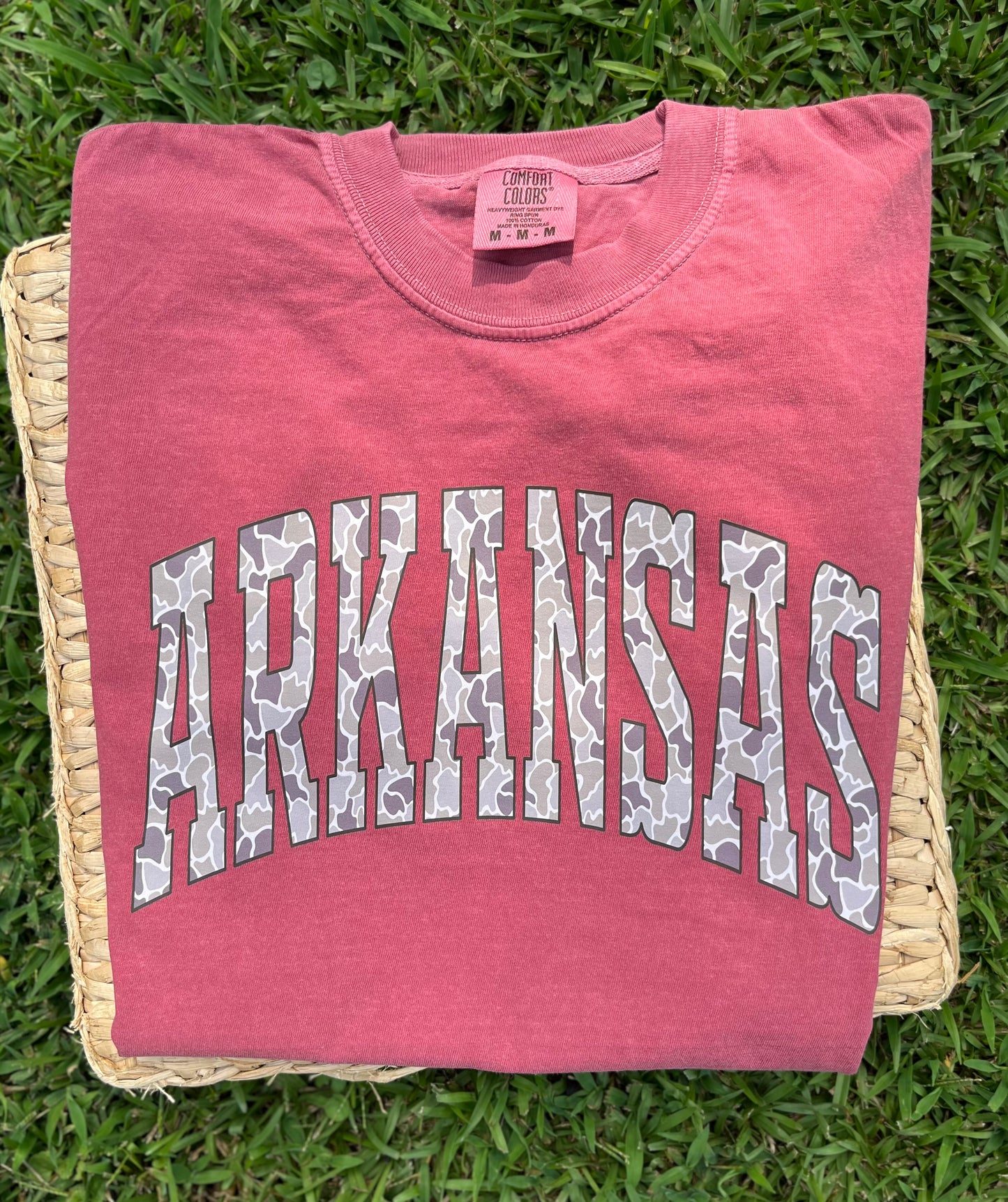 folded crimson red tee lying on a piece of wicker that is lying in the grass tee has the word arkansas in camo printed on front