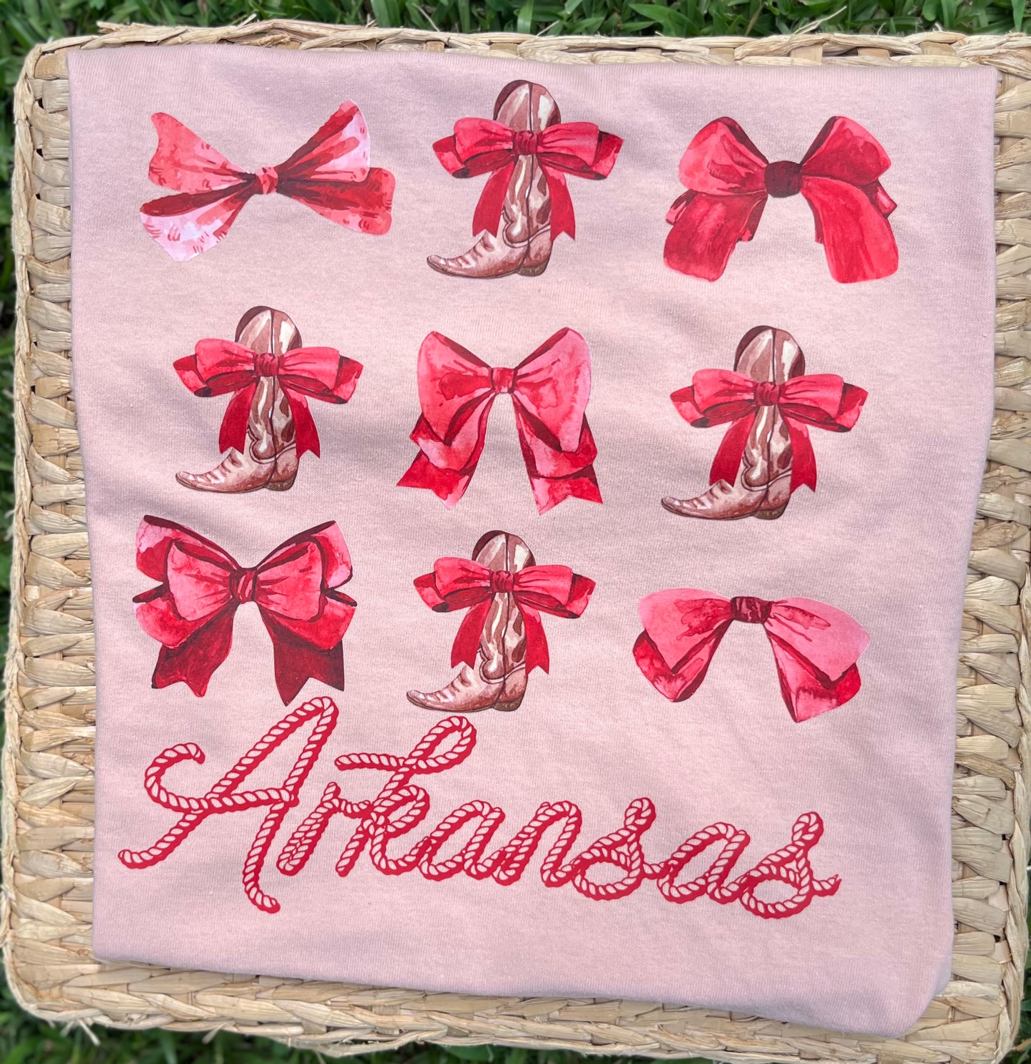 blush colored short sleeve tee, featuring a western-themed print with red bows, cowboy boots, and the word 'Arkansas' in red script, displayed on a woven mat over green grass.