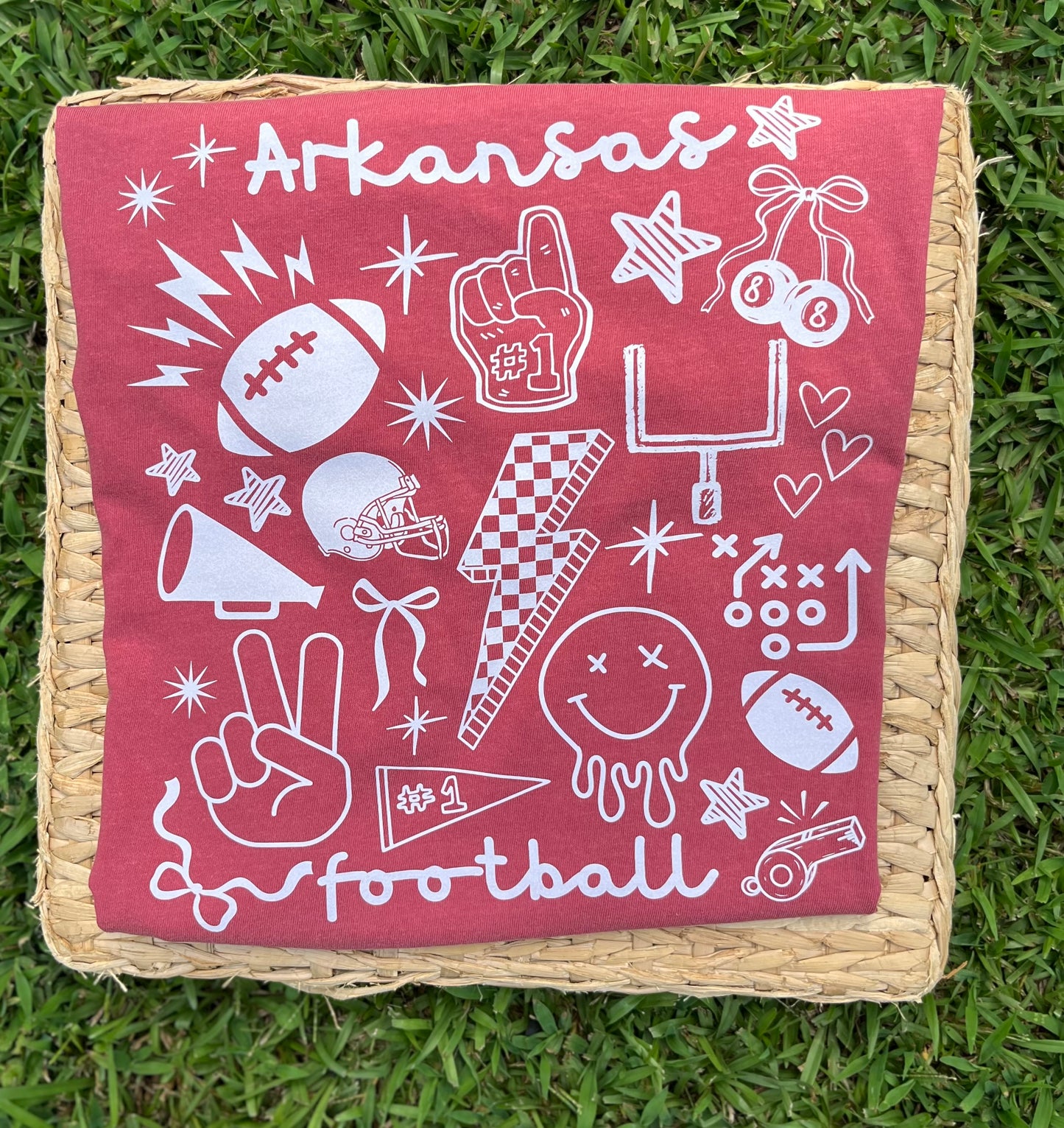 A square woven cushion with a crimson red short sleeve tee printed with white Arkansas football-themed graphics and text, placed on green grass.