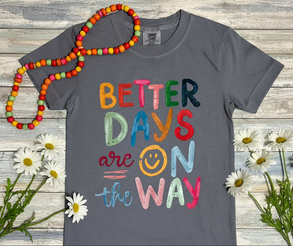 Gray t-shirt with colorful text 'Better Days are on the Way' with a smiley face in place of the letter "o" of the work "on" Pictured on a wooden surface with colorful bead stand and daisy flowers available in short or long sleeve tee