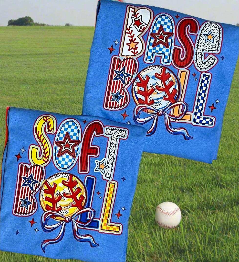 A blue t-shirt with a colorful baseball and softball design lying on a field with a baseball next to it.