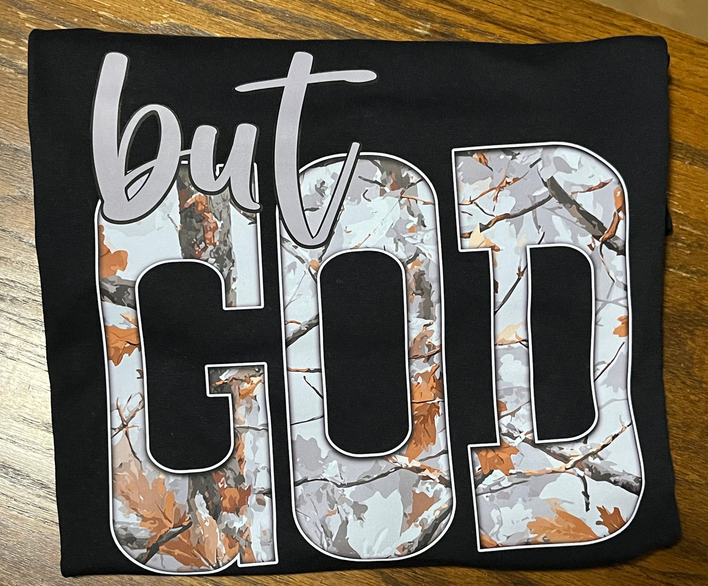 Black shirt with 'but God' printed in white and camouflage pattern on a wooden surface