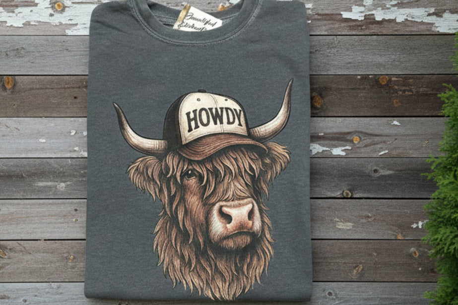 Gray tshirt with a brown highland cow wearing a 'Howdy' cap design on a wooden background
