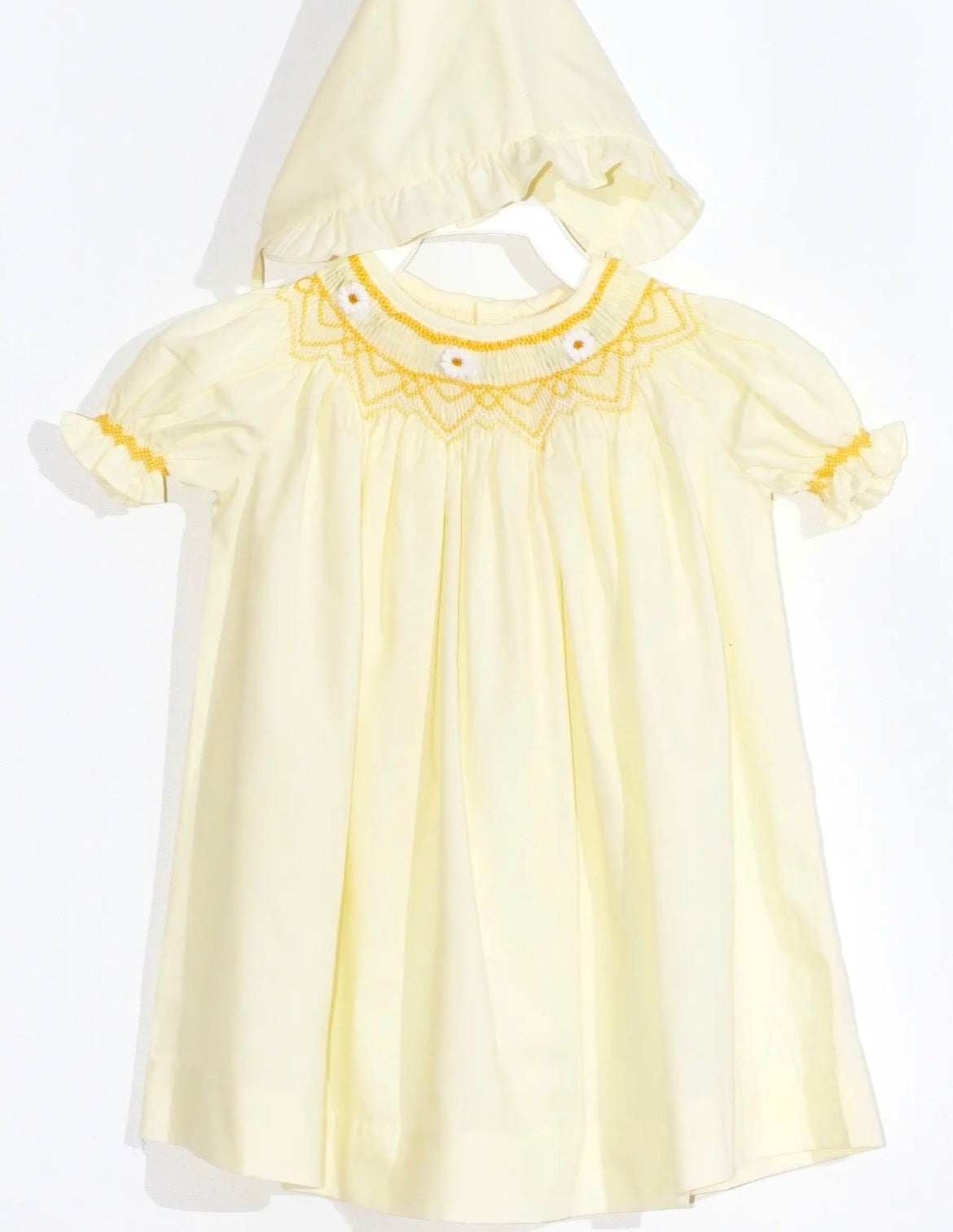 A pale yellow baby dress with a matching bonnet and bloomers featuring yellow embroidered detailing around the collar and small daisy flowers and yellow ruffle trim on the short sleeves, displayed on a white hanger against a white background.