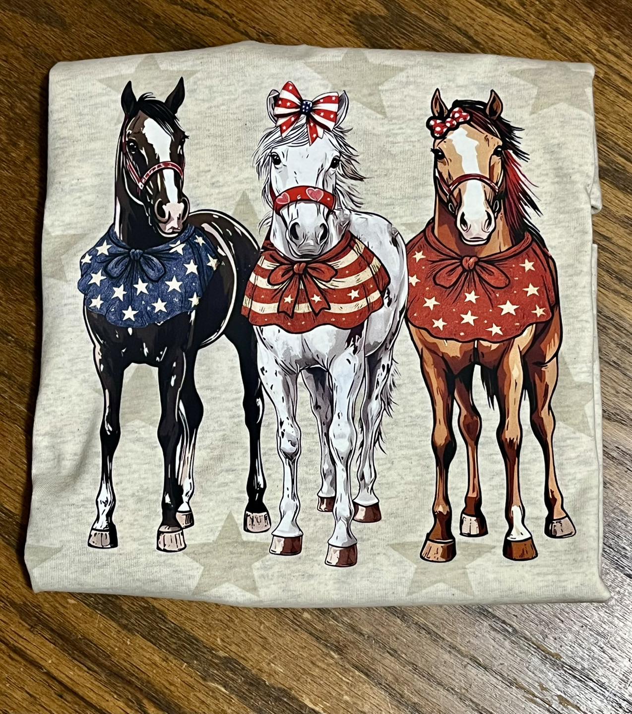 Folded tshirt with neutral color stars with three horses wearing American flag-themed bows on a wooden surface