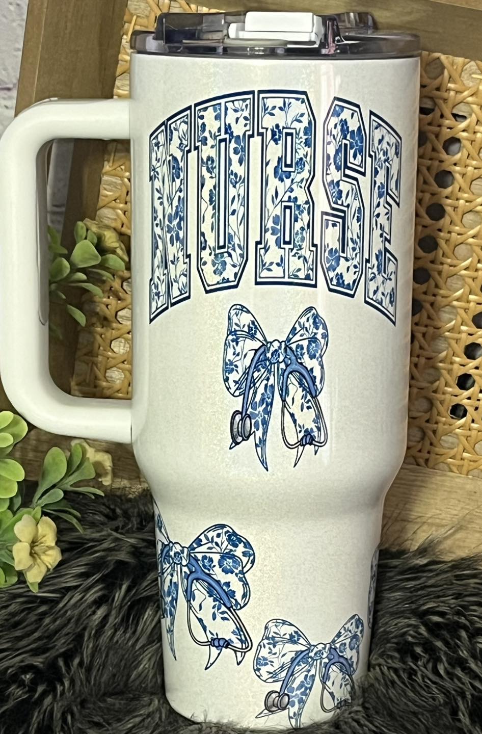 A 40oz with handle white stainless steel tumbler with blue floral designs and the word 'NURSE' displayed prominently. It includes a stethoscope bow decoration.