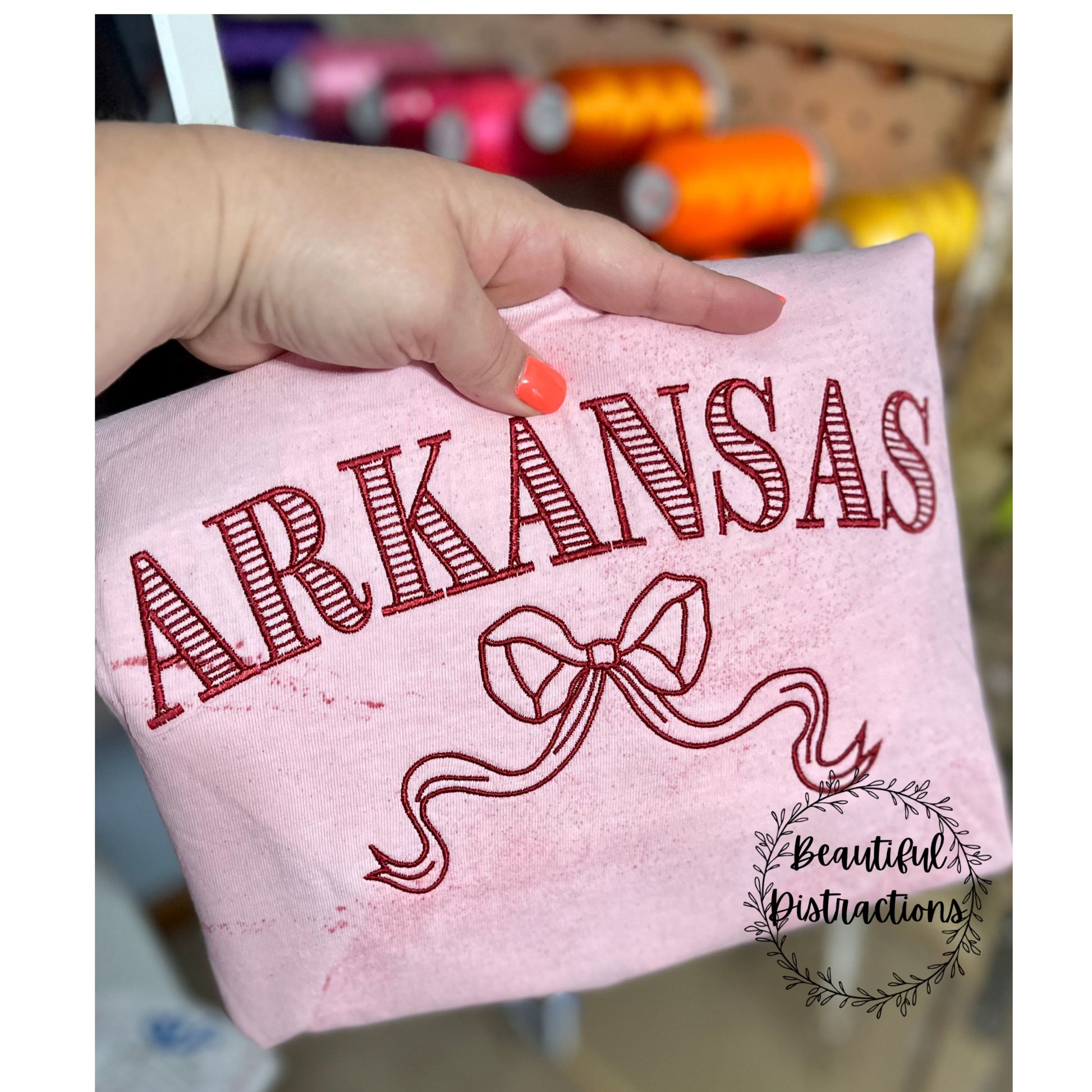 Comfort colors clay colorblast tee in unisex 100% cotton fit with 'ARKANSAS' and a bow embroidered design  in dark red thread, held by a hand.