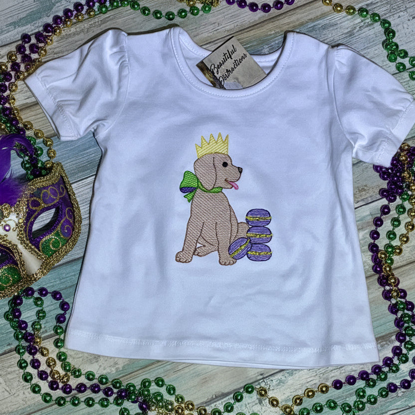 White t-shirt with a dog wearing a crown and bow, surrounded by Mardi Gras beads and mask on a wooden surface.