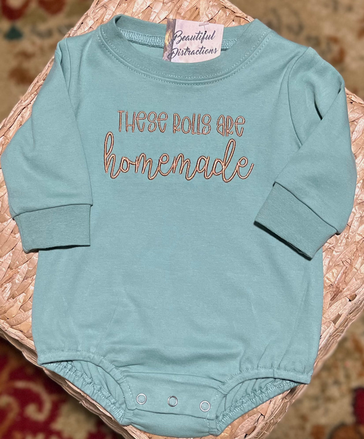 SAGE COLOR BABY BUBBLE ROMPER WITH THESE ROLLS ARE HOMEMADE EMBROIDERED ON THE FRONT CHEST AREA IN A BROWN THREAD.  LYING ON A WICKER SQUARE 