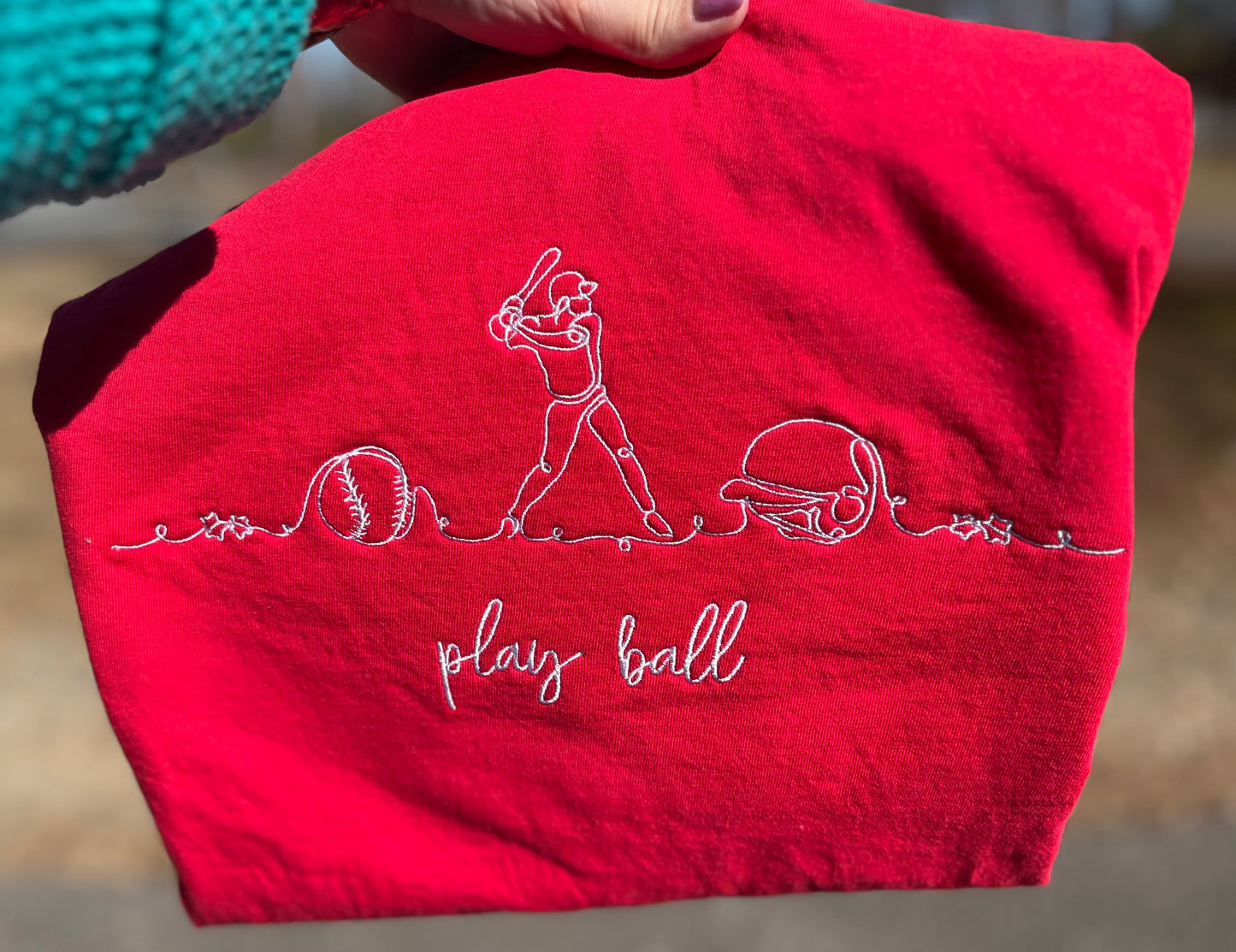 Red tshirt with white embroidered design of a baseball player, ball, helmet and stars and 'play ball' text.