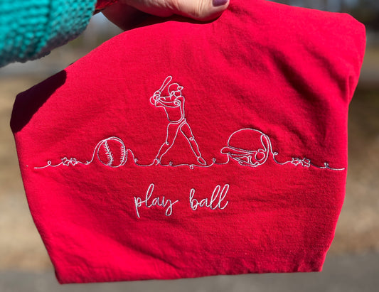 Red tshirt with white embroidered design of a baseball player, ball, helmet and stars and 'play ball' text.
