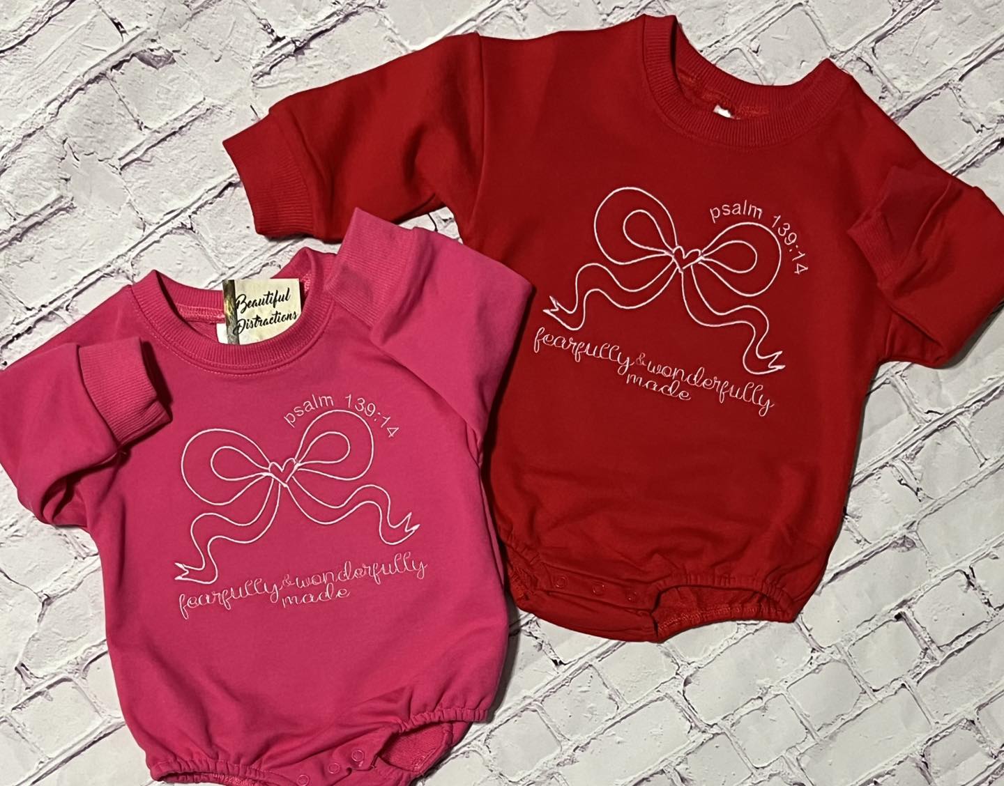 Two baby outfits, a pink romper and a red sweatshirt  romper, on a textured surface.
embroidered with fearfully and wonderfully made design that includes a bow with a heart in the middle