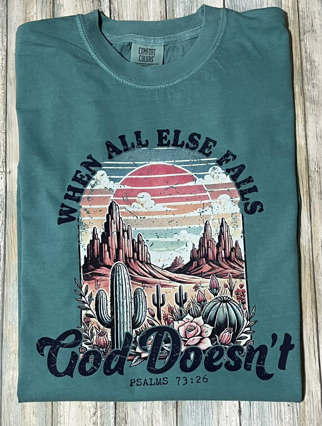 A green t-shirt with a graphic print of a desert landscape at sunset and the text 'God doesn't fail' from Psalms 71:12-16.