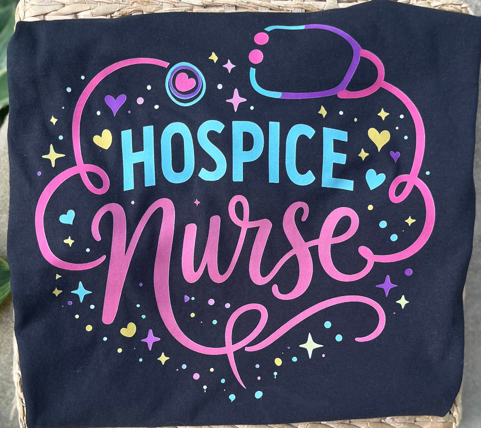 BLACK TEE WITH HOSPICE NURSE WORDING, HEARTS AND STETHOSCOPE IN A COLORFUL PRINT LYING ON A WOVEN SURFACE