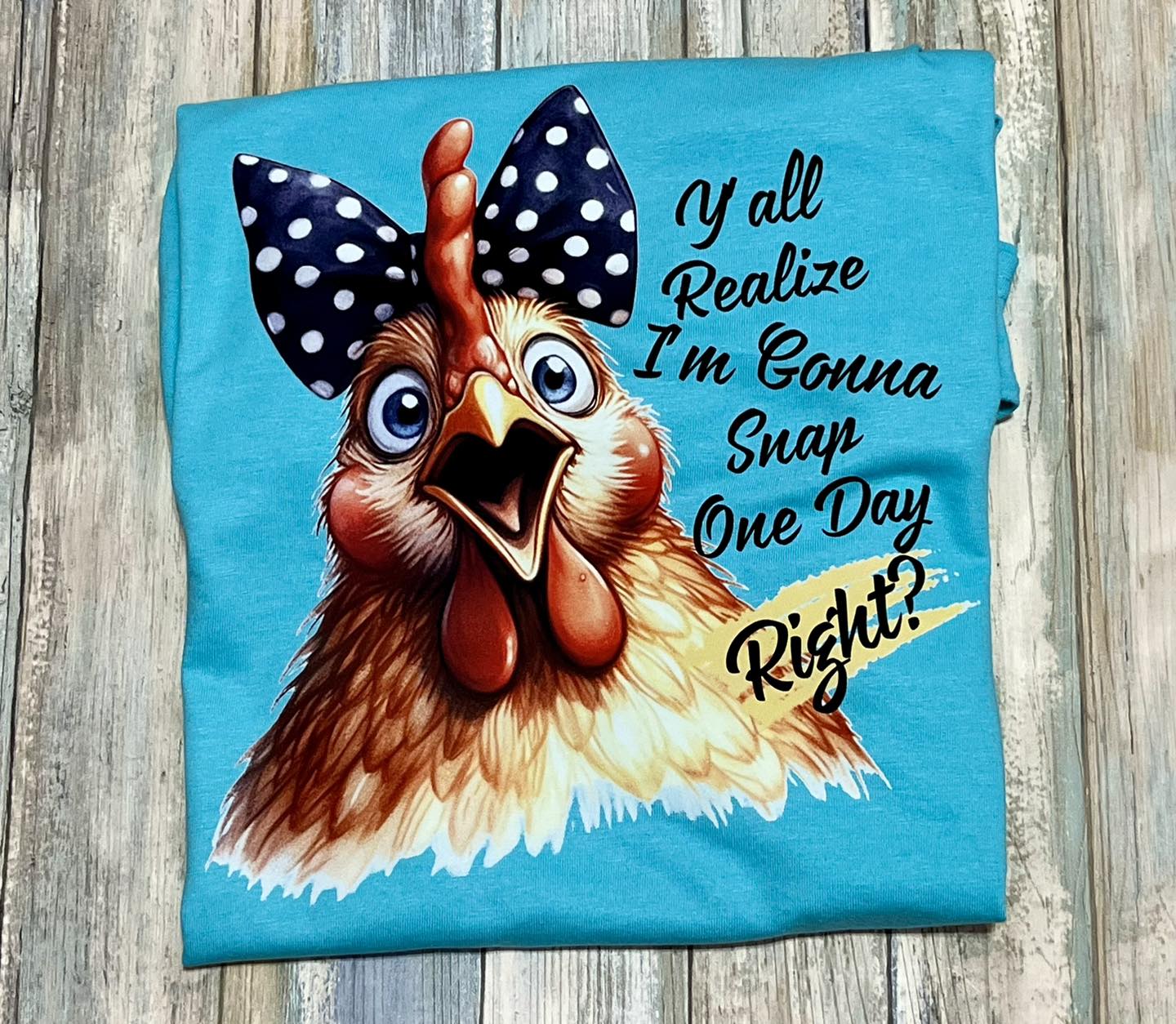 A blue t-shirt with a graphic print of a chicken wearing polka-dot hairbow and the text 'Y'all realize think I'm gonna snap one day right?'.