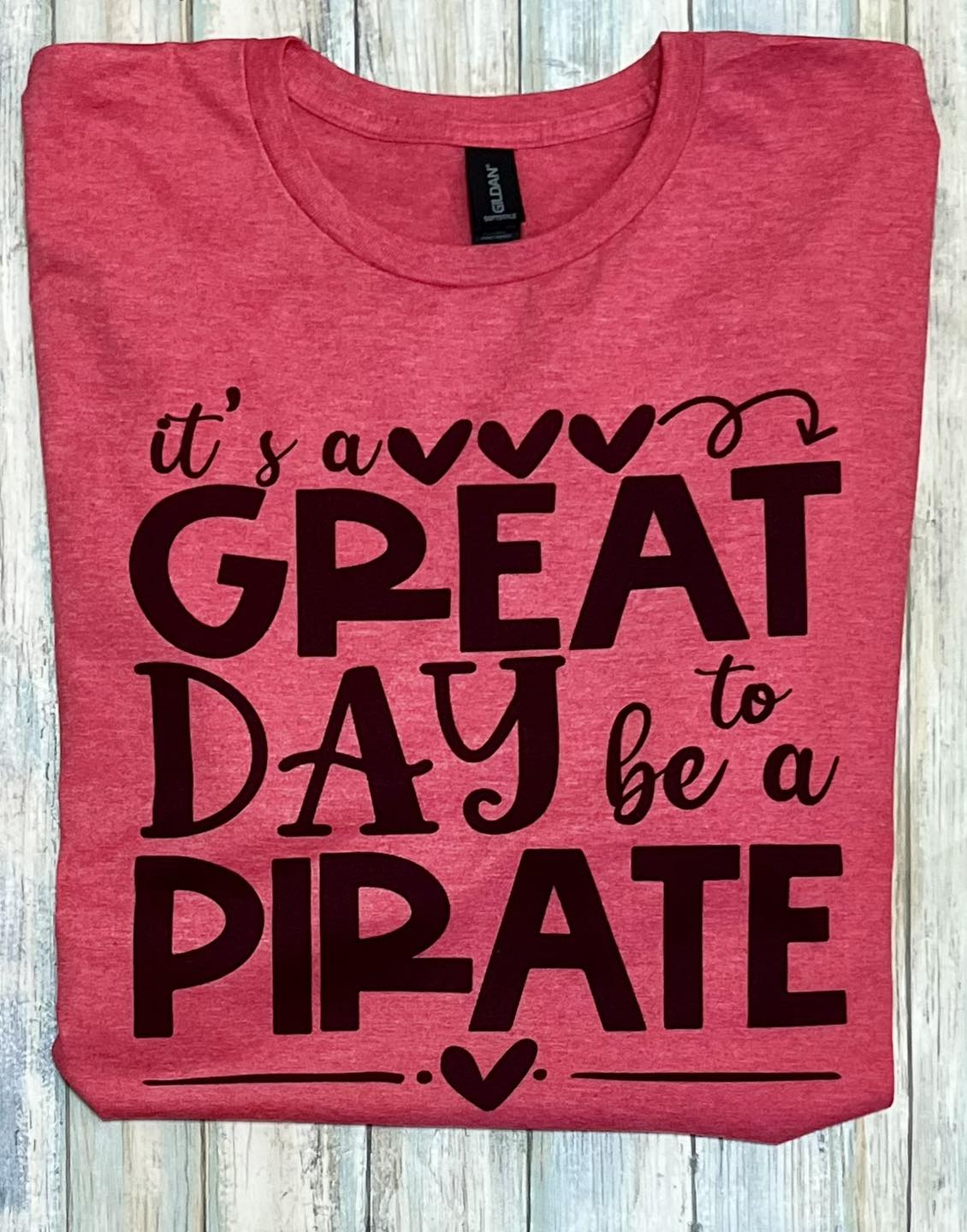 A heather red t-shirt with the text 'It's a GREAT DAY TO BE A PIRATE' printed in bold, cursive script, centered on the chest area.