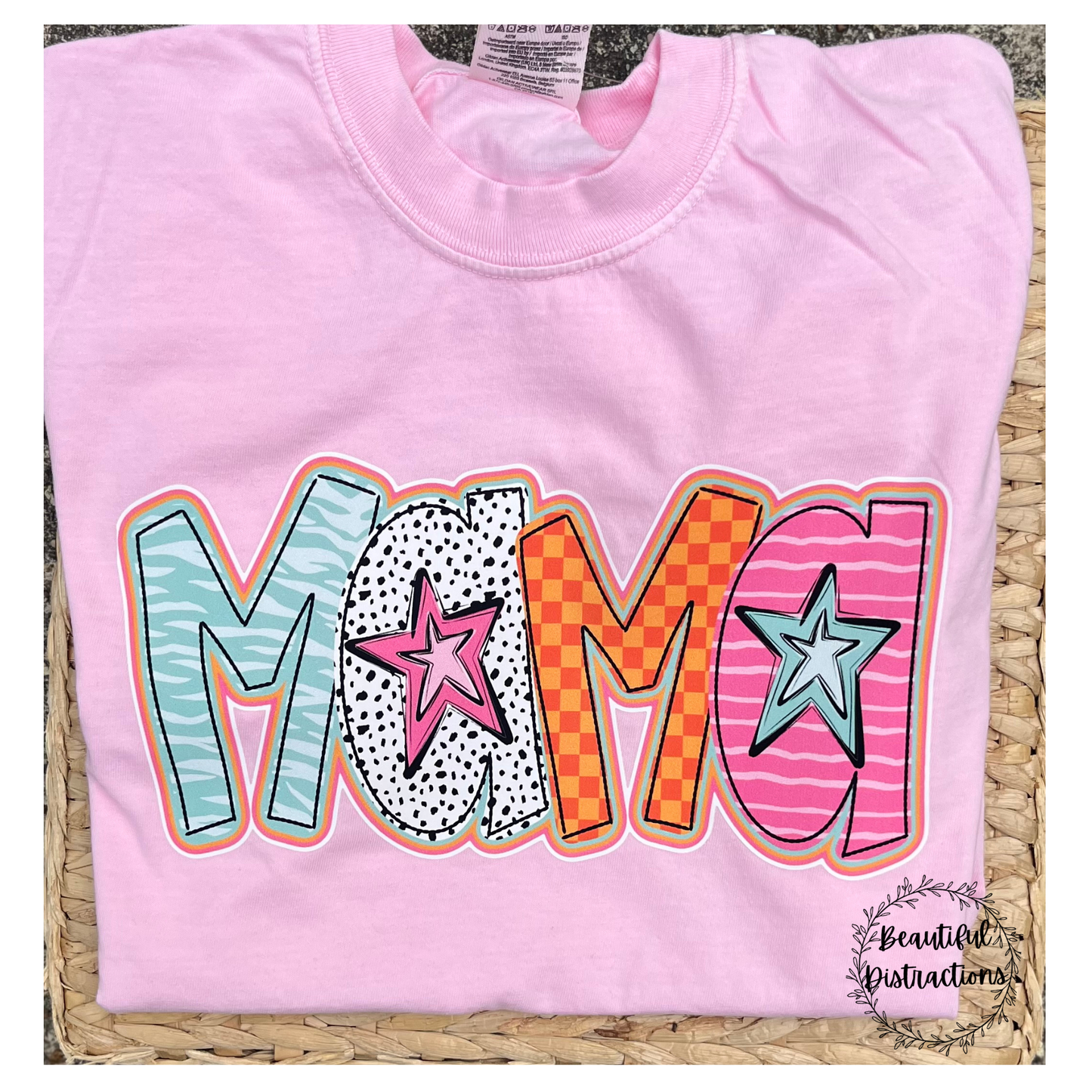 COLORFUL MAMA PRINT DESIGN IN DOODLE FONT WITH STARS  ON A PINK SHIRT LYING ON A WOVEN SURFACE 