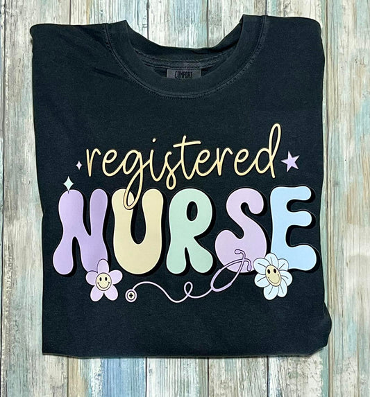 A pepper color t-shirt with the text 'REGISTERED NURSE' in a colorful, playful font, featuring illustrations of a smiling daisy and a cartoon-style flower.