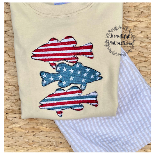 tan shirt with stacked embroidered stars and stripes fish in red white and blue. sitting on a pair of khaki and white striped shorts sitting on a whicker pad 