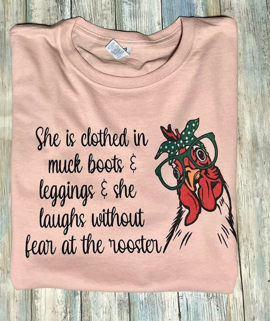 A blush color tshirt with an illustration of a chicken and the text phrase 'She is clothed in muck boots and leggings, laughing without fear at the rooster.'.