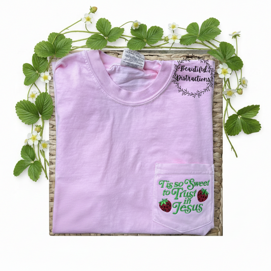 pick tee with tis so sweet to trust in Jesus and strawberries embroidered on the pocket in green.  lying on wicker background with strawberry vine lying around the top of the shirt