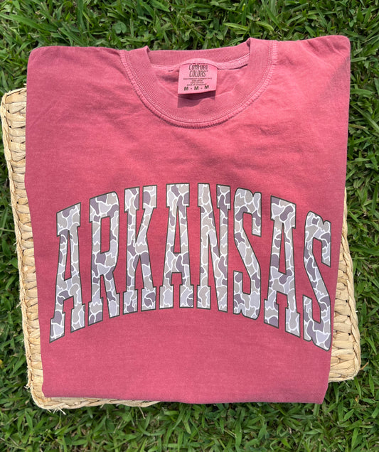 folded crimson red tee lying on a piece of  wicker that is lying in the grass   tee has the word arkansas in camo printed on front