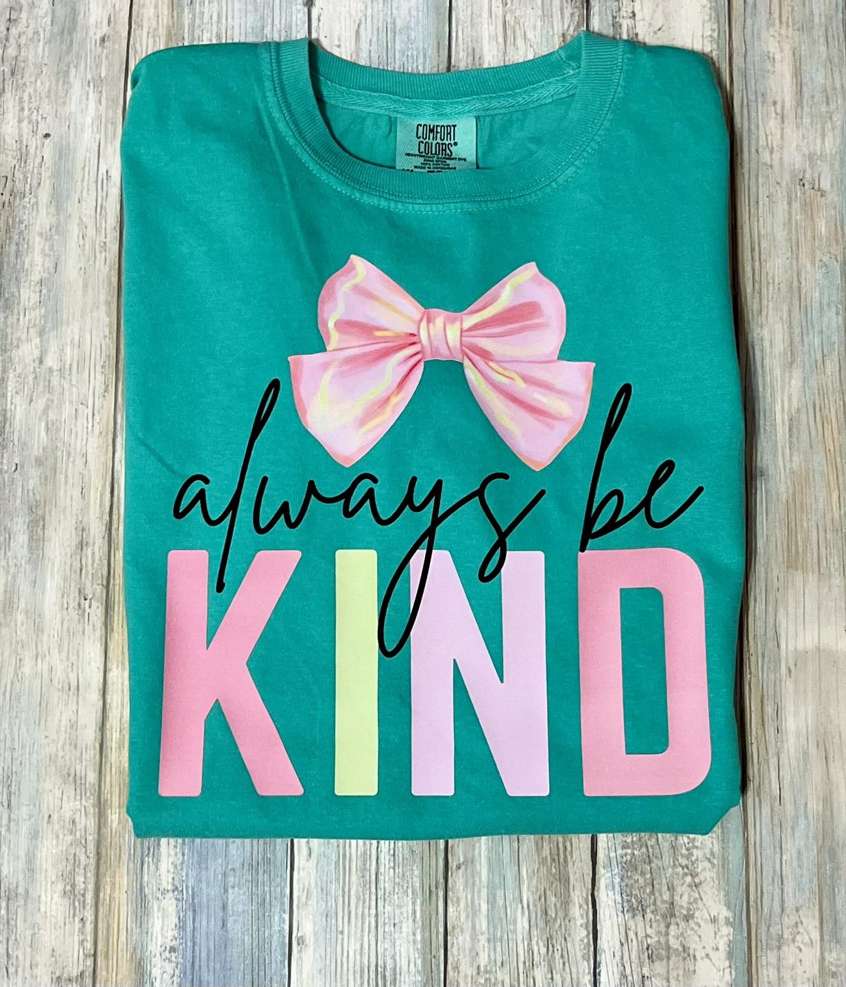 A folded green t-shirt with the words 'always be KIND' printed in a colorful, segmented font style. The phrase is accompanied by a pink  bow design.
