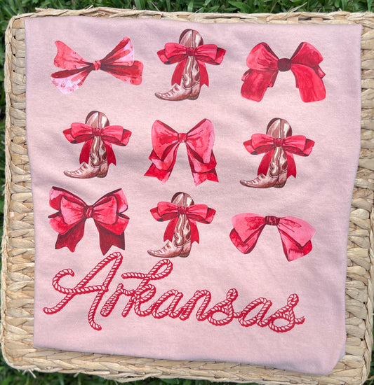 blush colored short sleeve tee, featuring a western-themed print with red bows, cowboy boots, and the word 'Arkansas' in red script, displayed on a woven mat over green grass.