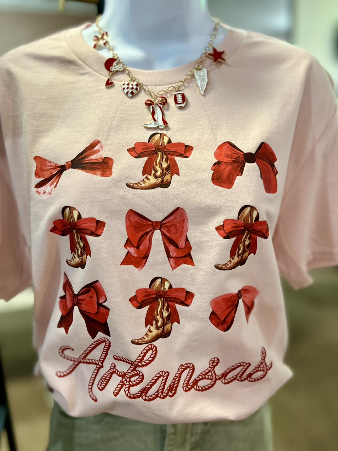 blush color t-shirt with red bows and brown boots design, featuring the word 'Arkansas', on a mannequin.