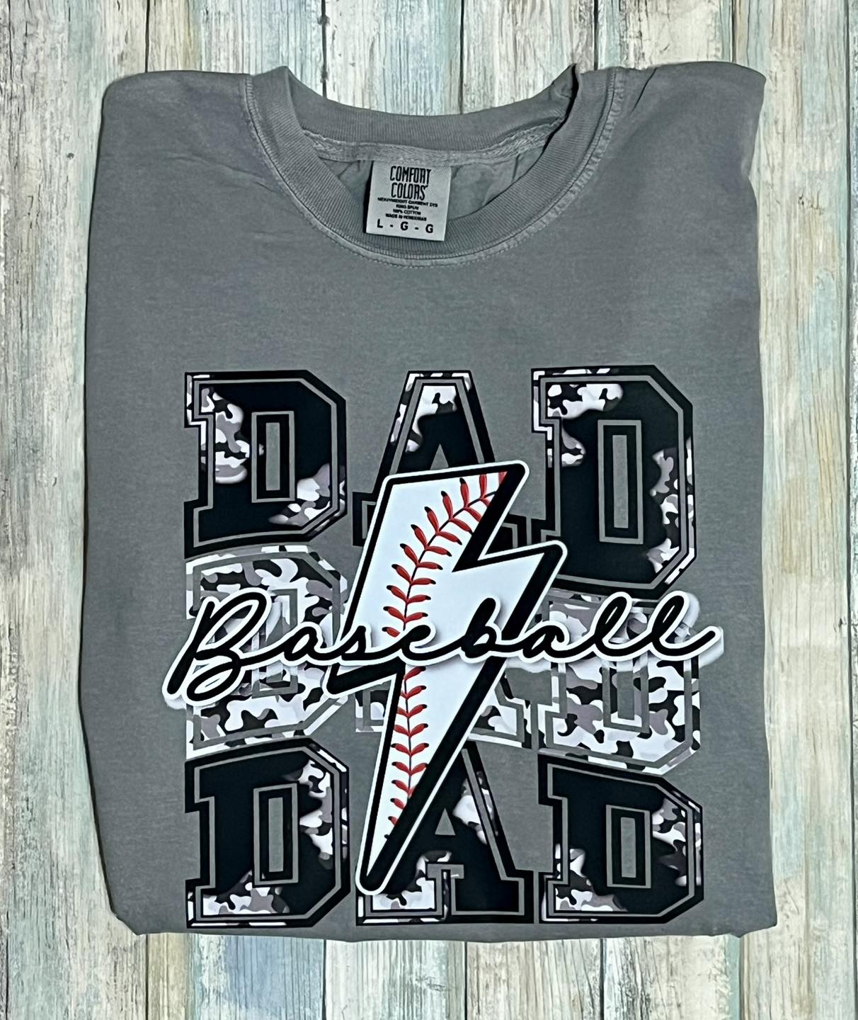 A gray t-shirt with gray and black camouflage pattern and text 'DAD Baseball' on it, displayed on a wooden surface.