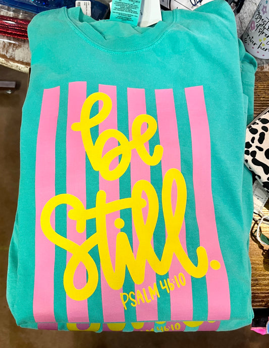 Teal shirt with pink stripes and yellow text 'be still'  and psalm 46:10 on a wooden surface.  available in long and short sleeve