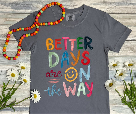 Gray t-shirt with colorful text 'Better Days are on the Way' with a smiley face in place of the letter "o" of the work "on"  Pictured on a wooden surface  with colorful bead stand and daisy flowers available in short or long sleeve tee