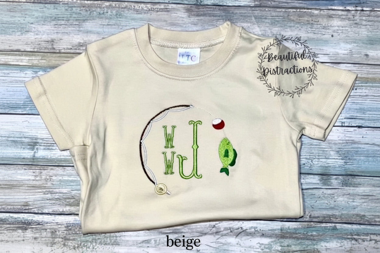 beige child shirt with fishing pole, bobber and fish embroidered on it and embroidered monogram in green in the middle  shirt is lying on a wooden surface