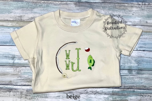 beige child shirt with fishing pole, bobber and fish embroidered on it and embroidered monogram in green in the middle  shirt is lying on a wooden surface