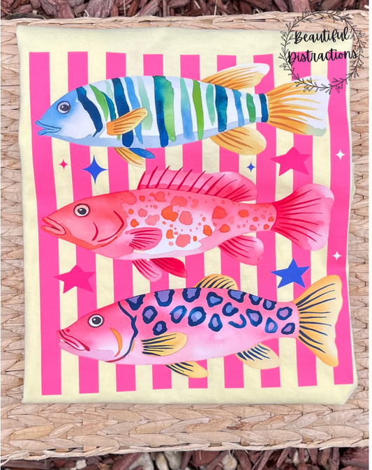 Colorful fish illustration on a pink striped background placed on a yellow tee.  shirt is placed on a wicker placemat with 'Beautiful Distractions' branding.
