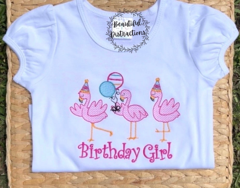 three flamingos embroidered on white shirt two wearing party hats and one holding two balloons  words birthday girl embroidered below them in pink  shirt is lying on woven flat lying on grass