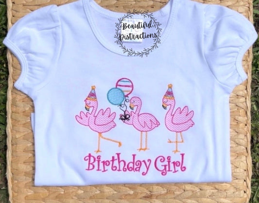 three flamingos embroidered on white shirt two wearing party hats and one holding two balloons  words birthday girl embroidered below them in pink  shirt is lying on woven flat lying on grass