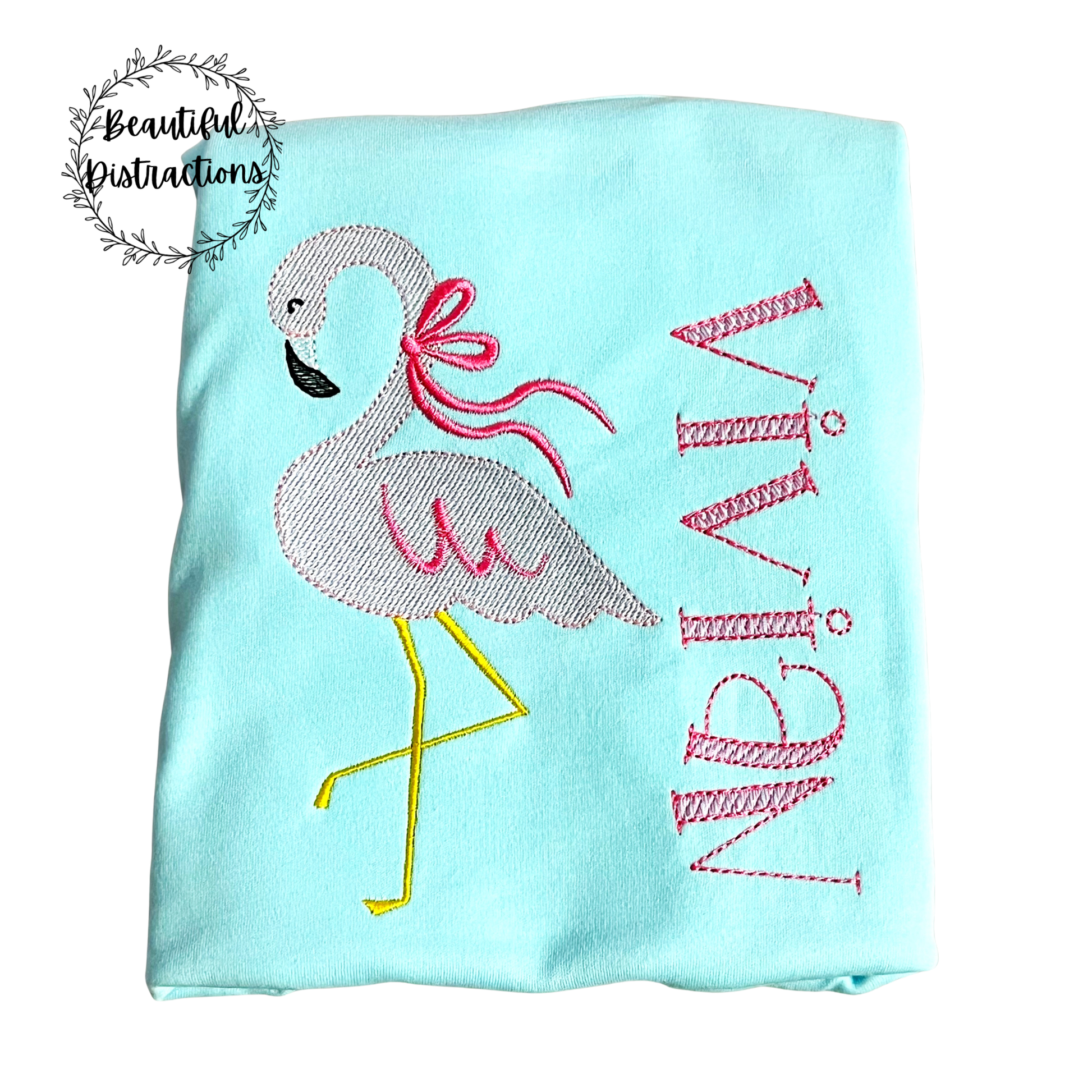 Light blue shirt with embroidered flamingo with bow on neck and vivian embroidered in pink lying on a white background