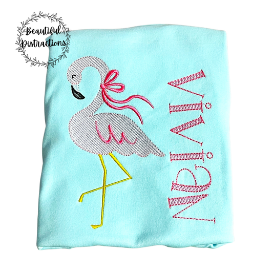 Light blue shirt with embroidered flamingo with bow on neck and vivian embroidered in pink lying on a white background