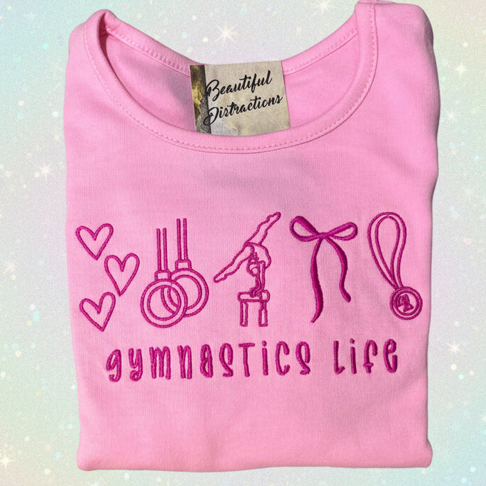 Pink folded shirt with embroidered gymnastics-themed design and 'gymnastics life' text on a glitter background.
