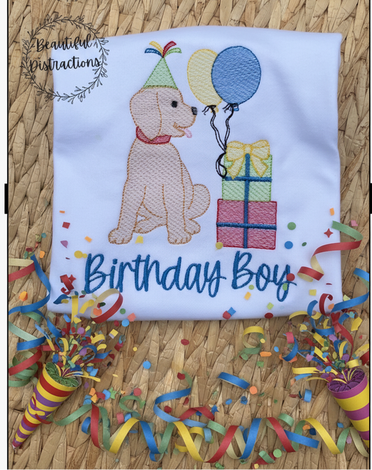 Embroidered shirt with a dog sitting beside balloons and wrapped girfts design and 'Birthday Boy' stitched below.  shirt sits on a woven surface and has party streamers lying around the bottom for decoration