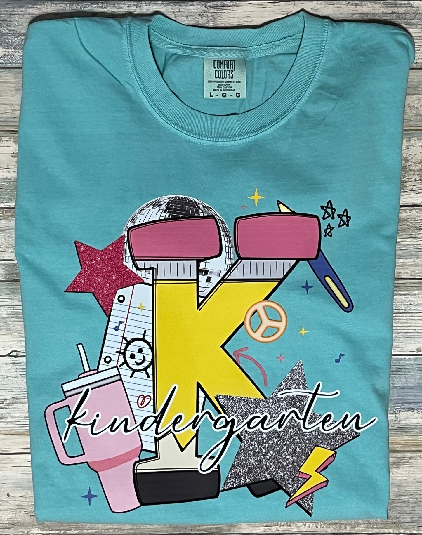 Teal t-shirt with colorful 'Kindergarten' design on a wooden surface
