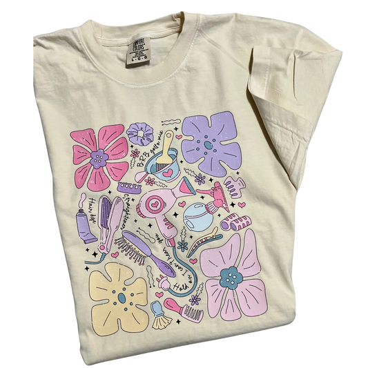A cream color t-shirt with a playful design featuring various hair styling tools and flowers, predominantly in pastel colors.