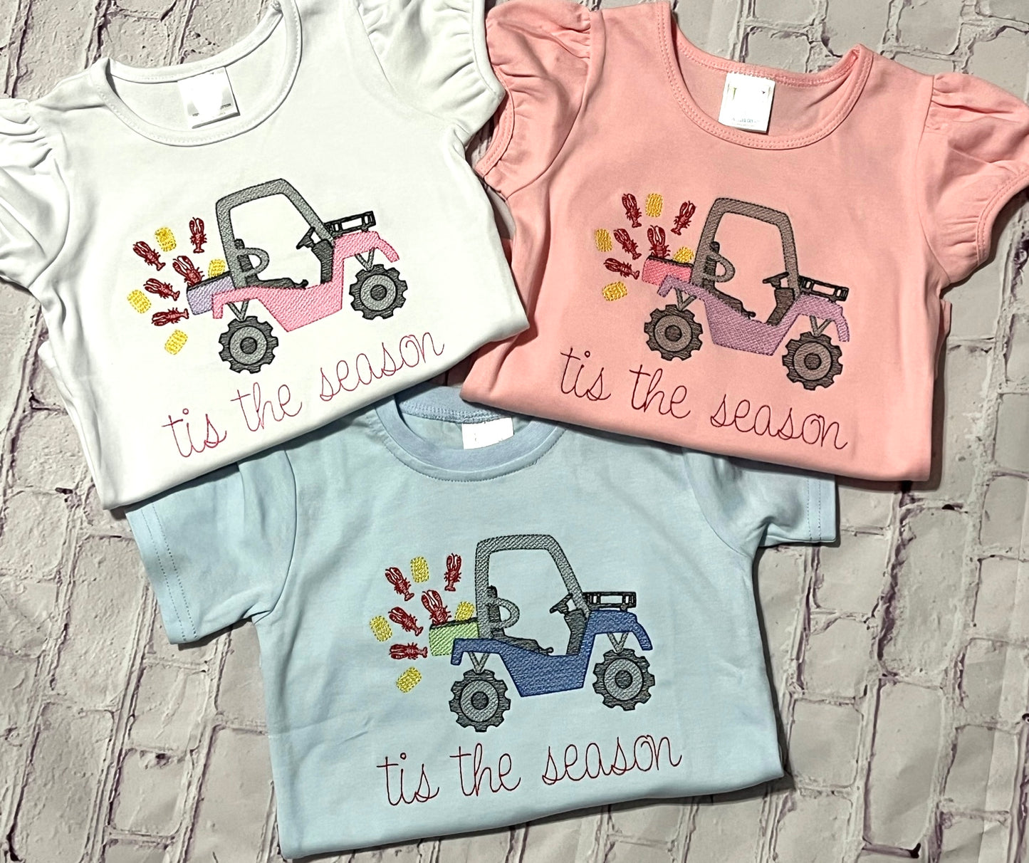Three children's t-shirts, one white, one pink and one blue with side by side and crawfish-themed designs on a stone surface.