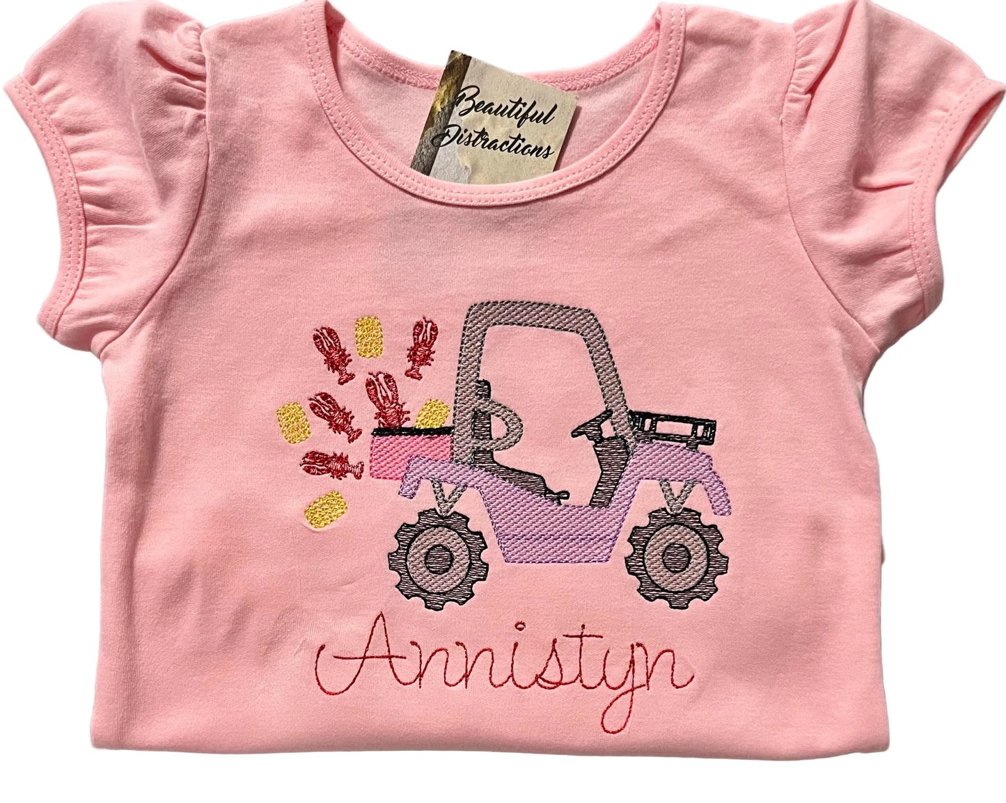 Pink t-shirt with a  side by side with crawfish and corn and embroidered name on a white background