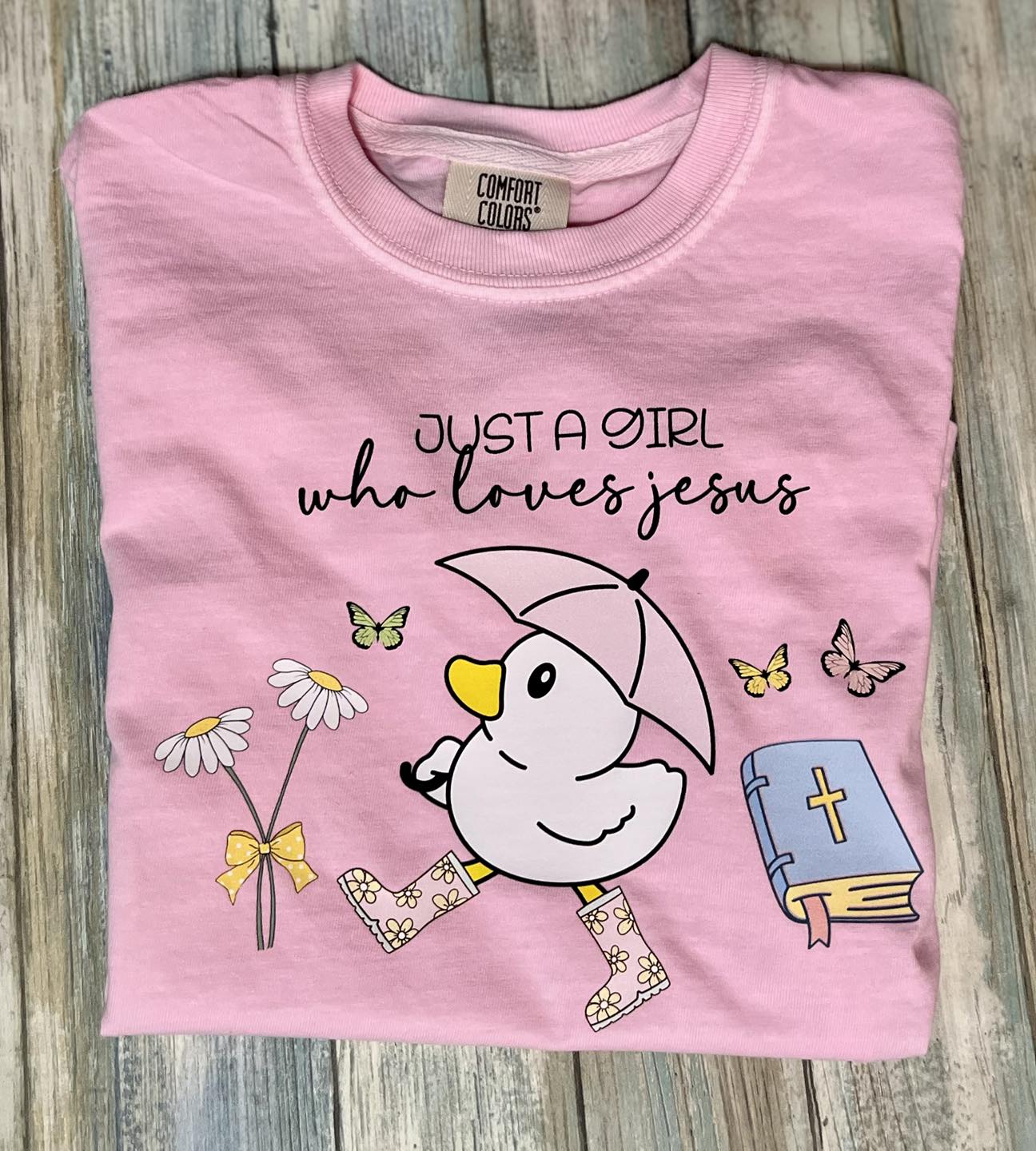 A pink t-shirt with the text 'JUST A GIRL who loves jesus' printed on the front, featuring an illustration of a duck, butterflies, and a book.