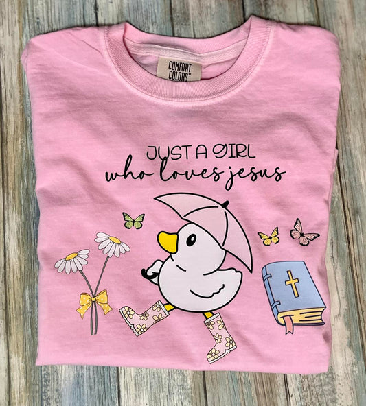 A pink t-shirt with the text 'JUST A GIRL who loves jesus' printed on the front, featuring an illustration of a duck, butterflies, and a book.