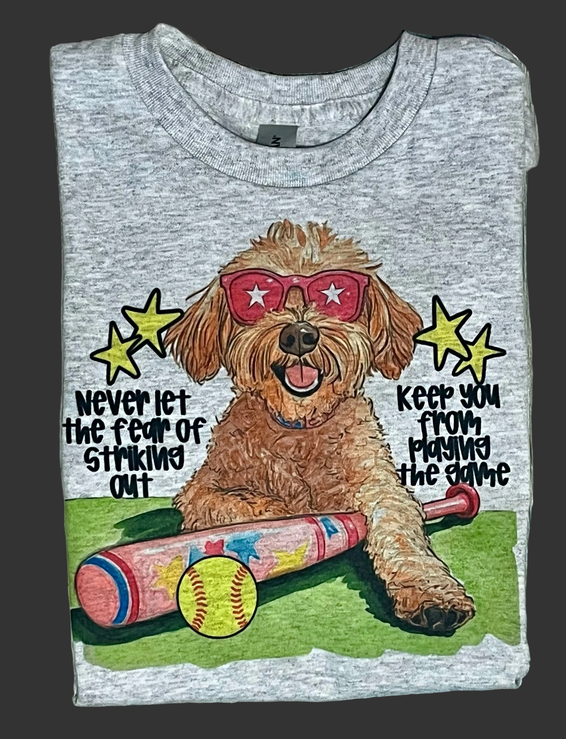 A graphic t-shirt with an illustration of a dog wearing sunglasses, a softball, and a softball bat, alongside text 'Never let the fear of striking out keep you from playing the game'.